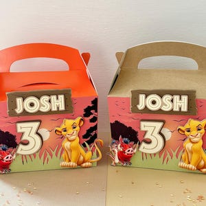 Puede incluir: Dos cajas de recuerdos de fiesta con asas, con un tema de El Rey León. Cada caja tiene el nombre "JOSH" y el número "3". Las cajas están decoradas con Simba, Pumbaa y un fondo de puesta de sol.