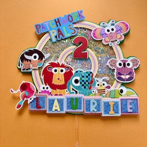 May include: A colorful cake topper with the text "PATCHWORK PALS" and the number "2". It features various cartoon animals, including a lion, koala, and butterfly, and the name "LAURIE" in denim-style letters. The background is orange.
