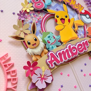 May include: A colorful Pokemon-themed cake topper featuring Pikachu, Eevee, and Bulbasaur. The topper includes the name "Amber" and the number "8", with decorative flowers and star-shaped confetti. The word "Pokemon" is also visible.