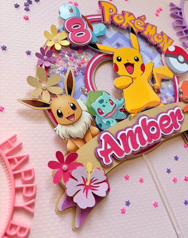 May include: A colorful Pokemon-themed cake topper featuring Pikachu, Eevee, and Bulbasaur. The topper includes the name "Amber" and the number "8", with decorative flowers and star-shaped confetti. The word "Pokemon" is also visible.
