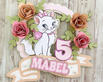 May include: A pink and white cake topper featuring a cartoon white cat with a pink bow, a pink number 5, and the name Mabel in pink letters. The topper is decorated with pink and orange paper flowers and green leaves.