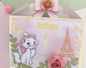 May include: A pink and white birthday party favor box with a gold trim. The box features a cartoon image of a white cat with a pink bow, a pink rose, and the Eiffel Tower. The text on the box reads "Bethany is 4".