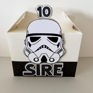 May include: A white party favour box featuring a black and white Stormtrooper helmet design. The box has the number "10" on top and the word "SIRE" on the front. The box is made of cardboard.