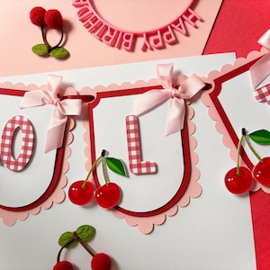 Peut inclure: Une guirlande festive avec le mot "LOL" épelé en lettres vichy rouges et blanches. Chaque lettre est sur une bannière blanche festonnée, ornée d'un nœud rose et d'une paire de cerises rouges. Une bannière "Happy Birthday" est en arrière-plan.
