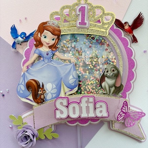 May include: A purple and gold cake topper featuring Sofia the First, a crown, a squirrel, a rabbit, a butterfly, and a bluebird. The topper has a glittery background and says "Sofia" on a banner.