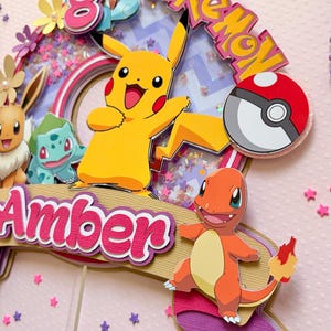 May include: A colorful Pokemon-themed cake topper featuring Pikachu, Charmander, and other characters. The topper includes the name "Amber" and the word "Pokemon" in a playful font, with a number 8. The background is pink with star-shaped confetti.