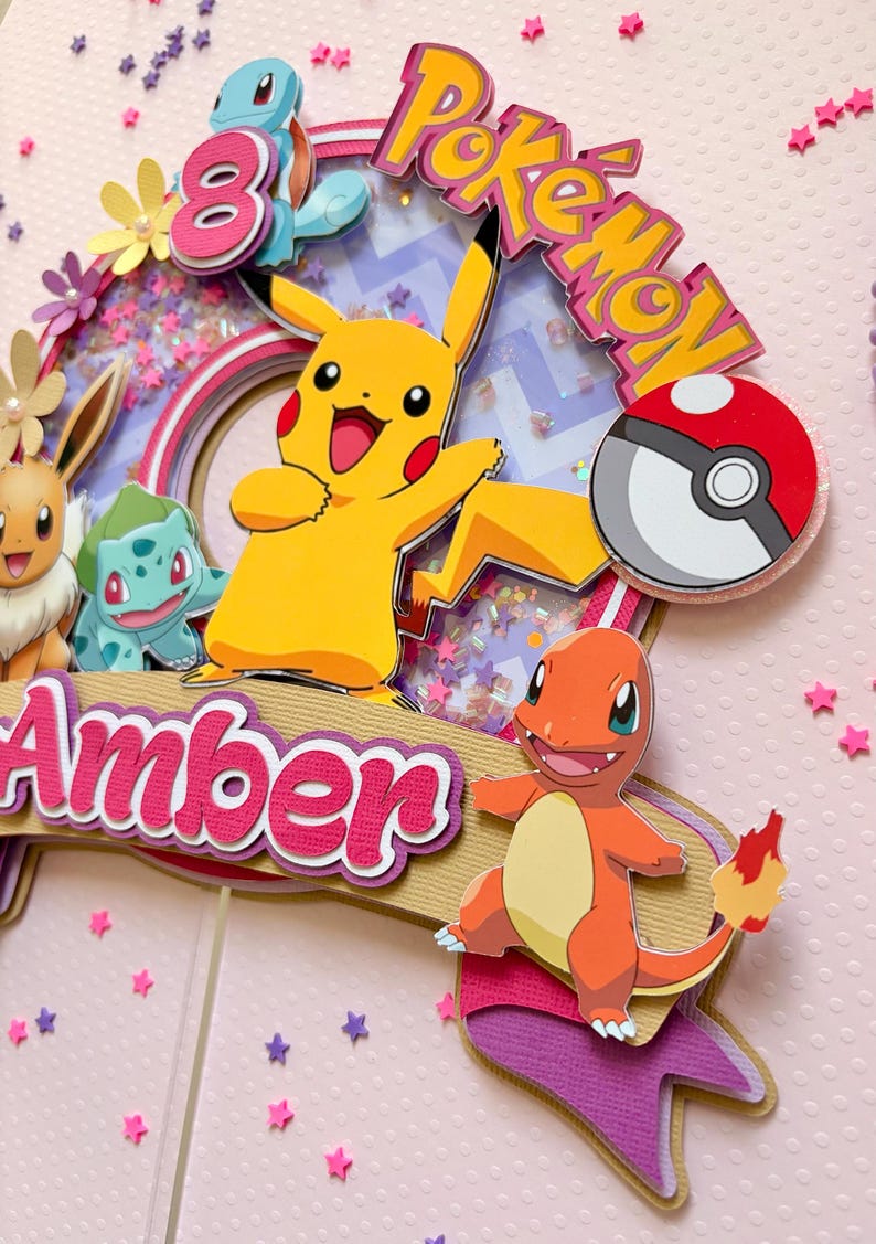 May include: A colorful Pokemon-themed cake topper featuring Pikachu, Charmander, and other characters. The topper includes the name "Amber" and the word "Pokemon" in a playful font, with a number 8. The background is pink with star-shaped confetti.