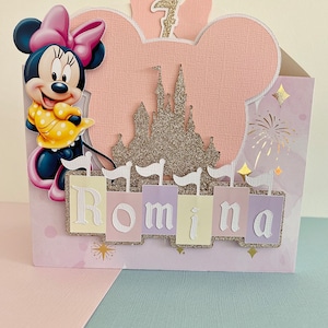 Może przedstawiać: R&oacute;żowa i biała kartka urodzinowa z motywem Minnie Mouse, złotym zamkiem z brokatem i imieniem Romina. Kartka ma r&oacute;żowe tło z białymi gwiazdami i fajerwerkami.