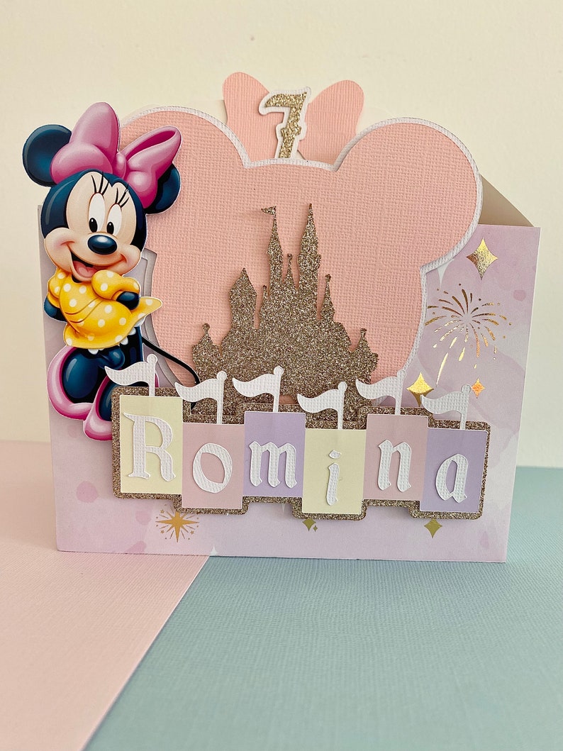Może przedstawiać: R&oacute;żowa i biała kartka urodzinowa z motywem Minnie Mouse, złotym zamkiem z brokatem i imieniem Romina. Kartka ma r&oacute;żowe tło z białymi gwiazdami i fajerwerkami.