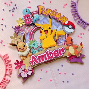 May include: A colorful Pok&eacute;mon-themed cake topper featuring Pikachu, Charmander, Squirtle, Bulbasaur, and Eevee. The topper includes the name "Amber" and the number "8". The design incorporates a Pok&eacute; Ball and the words "Happy Birthday".