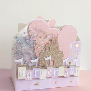 May include: A pink and white favour box with a castle, fireworks, and palm trees. The box has a pink Mickey Mouse ear design and the name "Harper-Ann" is spelled out on the front.