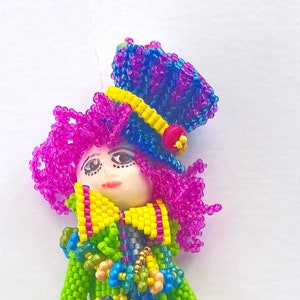 Puede incluir: Una mu&ntilde;eca de cuentas de colores con una peluca rosa, pantalones a rayas verdes y azules y un sombrero azul y morado. La mu&ntilde;eca lleva un lazo amarillo y tiene una chaqueta de cuentas verdes y azules. La mu&ntilde;eca est&aacute; de pie sobre un fondo blanco. Stephanieburnhamdesigns&copy;