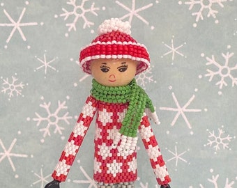 Puede incluir: Una muñeca de cuentas con un suéter rojo y blanco, una bufanda verde y un gorro blanco con un pompón. La muñeca lleva pantalones grises y patines de hielo negros. La muñeca está de pie sobre un fondo azul claro con copos de nieve blancos.