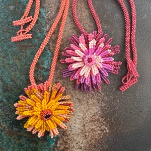 Pode incluir: Dois colares de contas com pingentes de flores. Os colares são feitos de pequenas contas de cor coral. Os pingentes são feitos de contas rosa, laranja e amarelas. Os pingentes têm a forma de flores com múltiplas camadas de pétalas.
