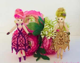 Puede incluir: Dos muñecas de cuentas con vestidos rosa y dorado se encuentran frente a flores rosas. Las muñecas tienen pelo de lana y caras pintadas. Las muñecas son hechas a mano y tienen un estilo caprichoso.
