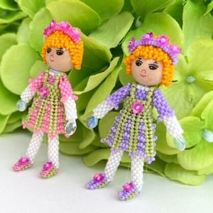 Pode incluir: Duas pequenas bonecas de contas com cabelo laranja, vestindo vestidos rosa e roxo com detalhes verdes. As bonecas usam sapatos brancos e coroas de flores.