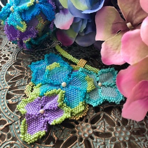 May include: A beaded flower bouquet with blue, green, and purple flowers. The flowers are made of small beads and are arranged in a cluster. The bouquet is sitting on a decorative metal tray.
