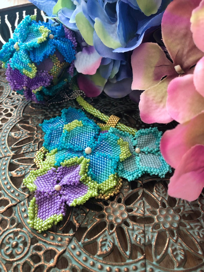 May include: A beaded flower bouquet with blue, green, and purple flowers. The flowers are made of small beads and are arranged in a cluster. The bouquet is sitting on a decorative metal tray.