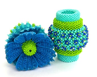 Puede incluir: Dos objetos decorativos de cuentas: una flor azul y un jarrón cilíndrico. La flor tiene pétalos azules y un centro verde. El jarrón es turquesa, verde y azul, con un estampado floral. Los artículos están hechos a mano y están sobre una superficie blanca.