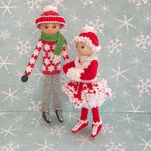 May include: Two beaded dolls wearing winter clothing and ice skates. The dolls are dressed in red, white, and green. The background is a light blue with a snowflake pattern.