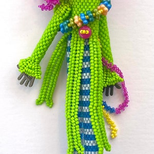 Puede incluir: Una mu&ntilde;eca de cuentas con un vestido a rayas verdes y azules, un sombrero rosa y azul y zapatos verdes. La mu&ntilde;eca tiene un bot&oacute;n en el pecho y una mano de plata.