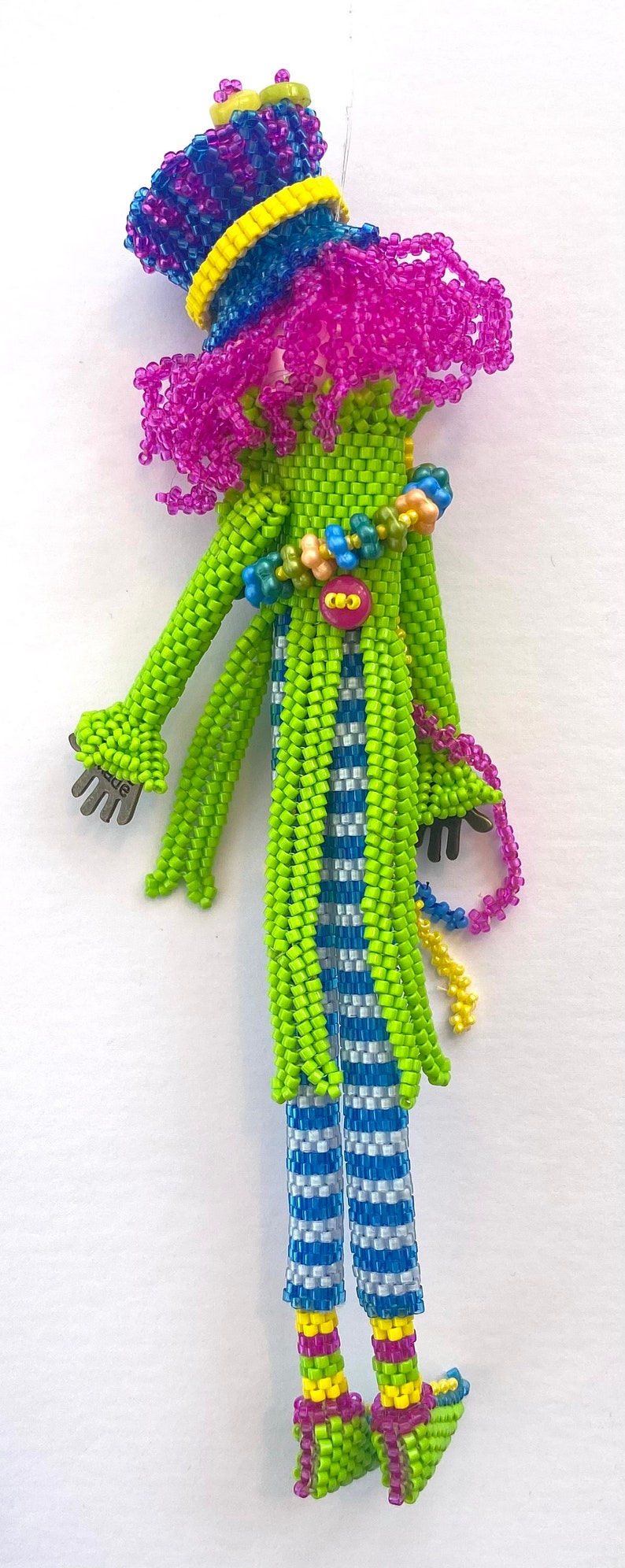 Puede incluir: Una mu&ntilde;eca de cuentas con un vestido a rayas verdes y azules, un sombrero rosa y azul y zapatos verdes. La mu&ntilde;eca tiene un bot&oacute;n en el pecho y una mano de plata.