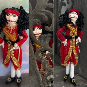 以下が含まれることがあります： ピーターパンのフック船長を模したビーズ人形。人形は赤と金の衣装を着ており、黒いベルトと黒い帽子をかぶっています。人形は右手にフックを持っています。