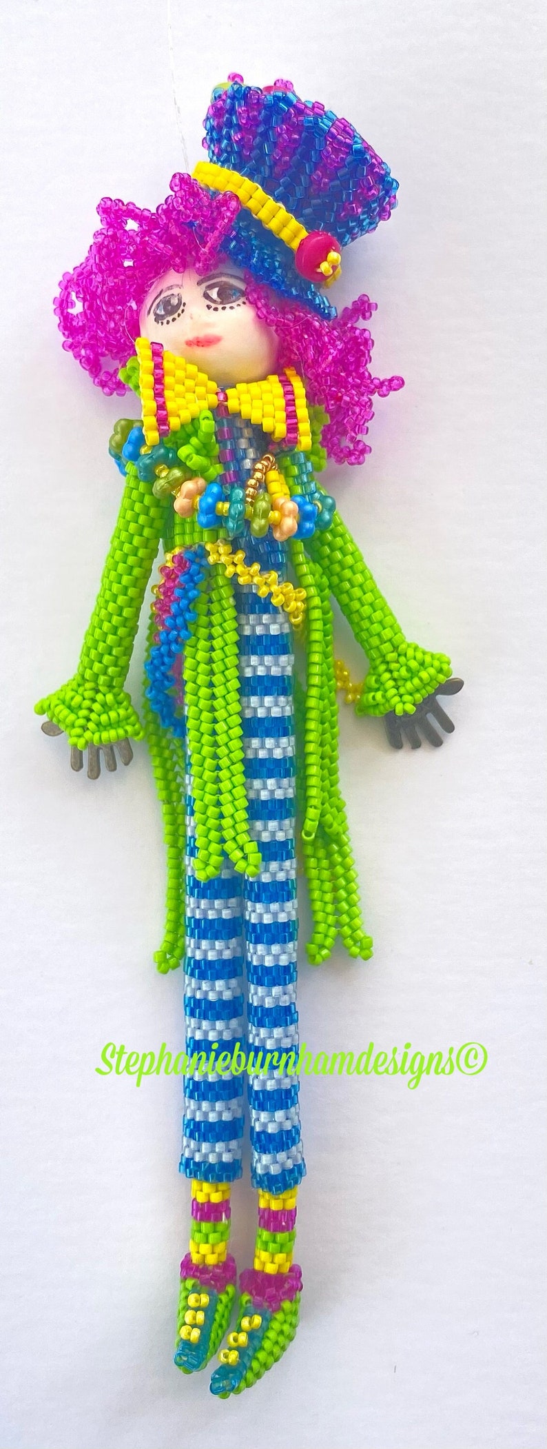 Puede incluir: Una mu&ntilde;eca de cuentas de colores con una peluca rosa, pantalones a rayas verdes y azules y un sombrero azul y morado. La mu&ntilde;eca lleva un lazo amarillo y tiene una chaqueta de cuentas verdes y azules. La mu&ntilde;eca est&aacute; de pie sobre un fondo blanco. Stephanieburnhamdesigns&copy;