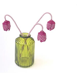 May include: A green ribbed glass vase holds three beaded flower stems. The stems are pink and white, with beaded flower buds in shades of pink and white. The vase is set against a white background.