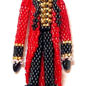 May include: A beaded doll wearing a red and black suit with a gold trim and a black top hat. The doll has a painted face with black eyes, a red mouth, and a black mustache.