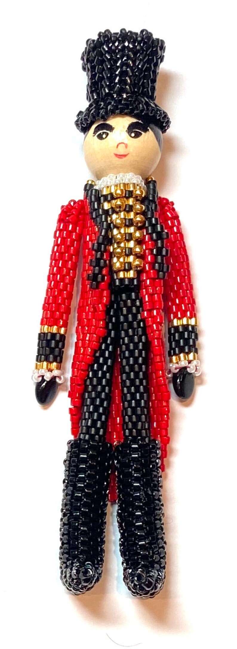 May include: A beaded doll wearing a red and black suit with a gold trim and a black top hat. The doll has a painted face with black eyes, a red mouth, and a black mustache.