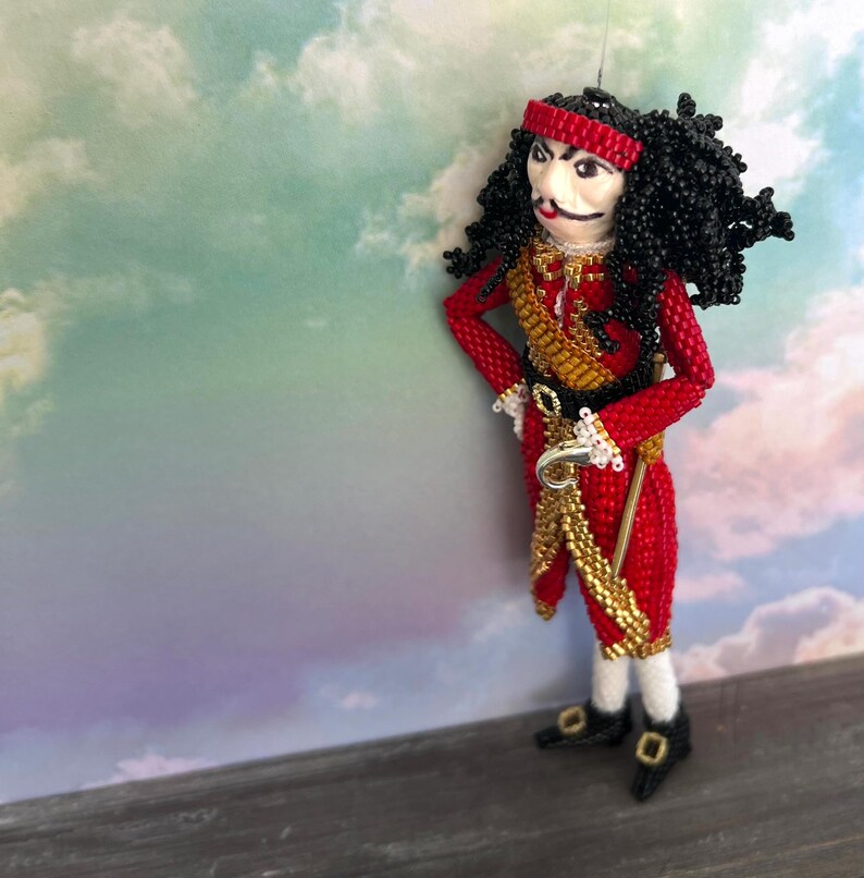 以下が含まれることがあります： 映画「ピーターパン」のフック船長をモチーフにしたビーズ人形。赤い金色の衣装を着て、黒いカツラをかぶり、手にフックを持っている。