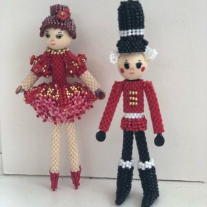May include: A red and white beaded nutcracker doll stands next to a red and pink beaded ballerina doll. Both dolls have painted faces and black hair.