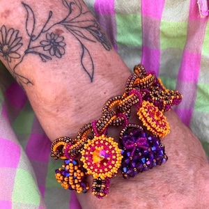 Könnte beinhalten: Ein Perlen-Charm-Armband mit einer Kette aus metallisch bronzefarbenen Saatperlen. Das Armband enthält mehrere hängende Anhänger, darunter einen runden Anhänger mit einem zentralen orangefarbenen Kristall, ein violettes Perlenherz und weitere Formen.