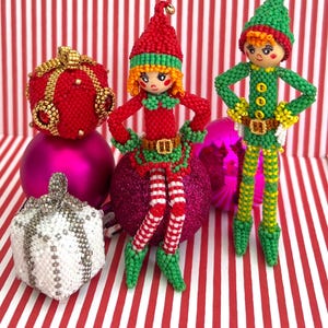 May include: Handmade Christmas decorations featuring beaded elf figures in red and green outfits, with matching hats and shoes. The ornaments are posed with their hands on their hips. Other decorations include a red beaded gift and a silver and white beaded ornament.
