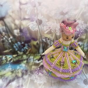 May include: A small doll with a pink and purple dress and a crown of pink flowers. The doll is made of beads and has a beaded skirt with butterflies. The doll is standing on a floral background.