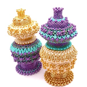 May include: Two beaded decorative objects. One is gold and cream, the other is purple, teal, and gold. Both have a bulbous shape with a crown-like top. The objects are intricately beaded with small, round beads.