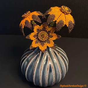 May include: A beaded floral sculpture featuring three yellow and metallic bronze flowers with central crystals, arranged in a ribbed, blue-grey ceramic vase. The text StephanieBurnhamDesigns is visible in the bottom right corner.