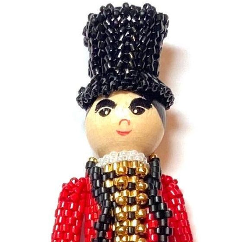 May include: A beaded doll wearing a black top hat, red jacket, and black and gold vest. The doll has a painted wooden face with a smile.
