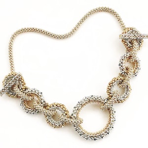 Könnte beinhalten: Eine silberne und goldene Perlenkette mit einem Kettenglied-Design. Die Kette besteht aus kleinen, runden Perlen, die miteinander verwoben sind, um ein zartes und kompliziertes Muster zu schaffen.