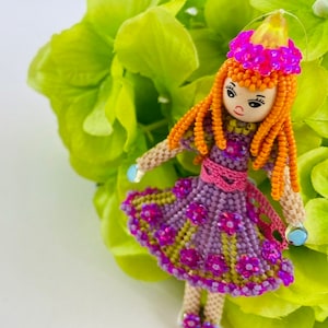 May include: A beaded doll with orange hair, a purple dress with pink flowers, and a pink crown. The doll is wearing a pink sash and has a light blue bead on each hand.