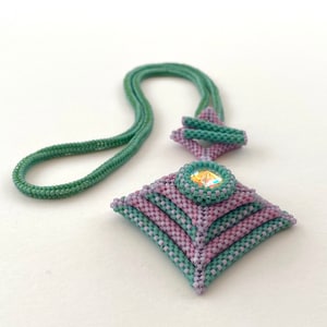 Puede incluir: Un collar de cuentas verde y morado con un colgante cuadrado que presenta un cristal transparente en el centro.