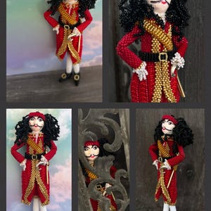 以下が含まれることがあります： 映画「ピーターパン」のフック船長をモチーフにしたビーズ人形。赤い金色の衣装を着て、黒い帽子と黒い髪をしています。人形は右手に剣を持っています。