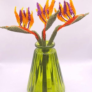 May include: A beaded floral arrangement featuring three Bird of Paradise flowers in a green glass vase. The flowers have orange, yellow, and purple beaded petals with green stems. The vase is a light green color.