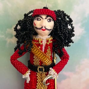 Pode incluir: Uma figurinha de contas do Capitão Gancho de Peter Pan. A figurinha está vestindo um traje vermelho e dourado com um cinto preto e um gancho na mão. A figurinha tem cabelo preto e um chapéu vermelho.