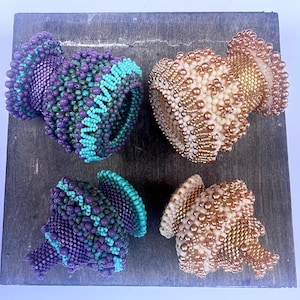 May include: Four beaded decorative objects in shades of purple, teal, and gold. The objects are vase-shaped, with intricate beadwork creating textured surfaces. The objects are arranged on a dark gray wooden surface.