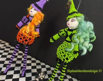 Puede incluir: Dos adornos de bruja de Halloween hechos con cuentas, con piernas largas, uno con ropa morada y naranja y cabello naranja, el otro con ropa verde y negra y cabello verde. Los adornos cuelgan de una cuerda blanca.