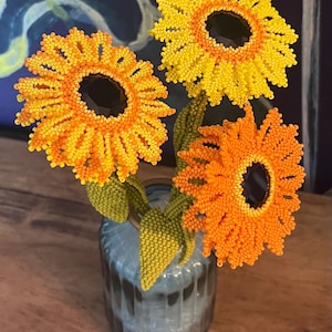 May include: Three handcrafted beaded flowers with yellow and orange petals and dark centers, featuring green beaded stems and leaves, arranged in a small, ribbed, translucent glass vase. The flowers are constructed from tiny seed beads.