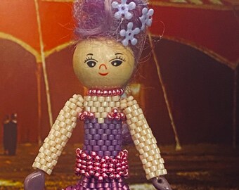Puede incluir: Una muñeca de cuentas de color morado y rosa con una peluca morada y pinzas para el pelo con flores moradas. La muñeca lleva un vestido morado con un ribete de cuentas rosas. La muñeca está de pie frente a una carpa de circo de rayas rojas y blancas.