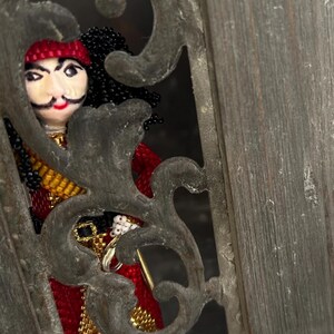 以下が含まれることがあります： 装飾的な金属製の枠から顔をのぞかせる、赤い帽子をかぶった黒髪のビーズ製の海賊人形。